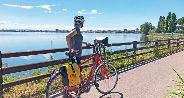 Radfahrer steht mit rotem Fahrrad an einem ruhigen Seeuferweg an einem klaren sonnigen Tag.