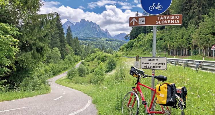 Rotes Fahrrad lehnt gegen Straßenschild, das nach Tarvisio Slowenien zeigt, mit zerklüfteten Bergen und Kiefernwald-Landschaft.