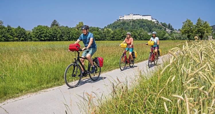 Drei Radfahrer fahren auf einem ländlichen asphaltierten Weg, der von grünen Feldern gesäumt ist, während eine Burg auf einem Hügel die Kulisse des blauen Himmels dominiert.