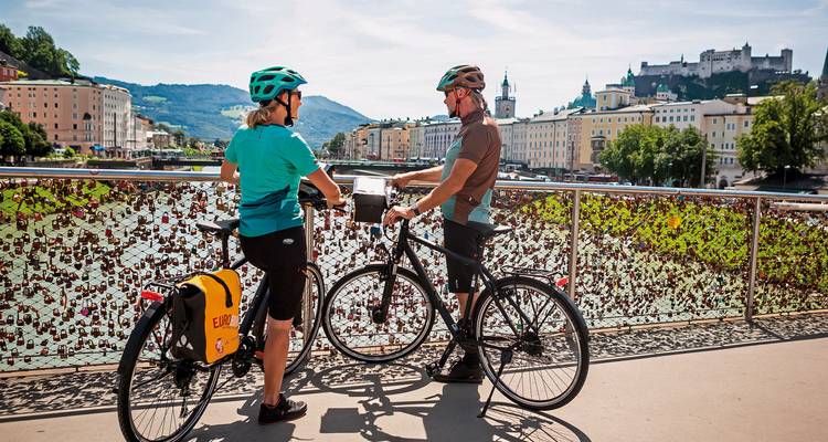 Zwei Radfahrer machen eine Pause auf einer mit Liebesschlössern bedeckten Brücke, während Salzburgs Altstadt und Festung vor einer Bergkulisse aufragen.