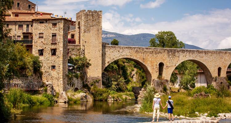 Dos personas están paradas cerca de un puente de piedra sobre un río con un telón de fondo de colinas y edificios históricos.