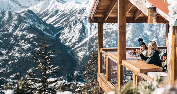 Une vue sur une montagne enneigée depuis un balcon en bois avec deux personnes savourant des boissons.