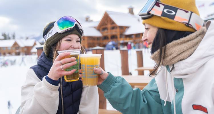 Deux femmes en tenue de ski avec des boissons, se souriant l'une à l'autre.