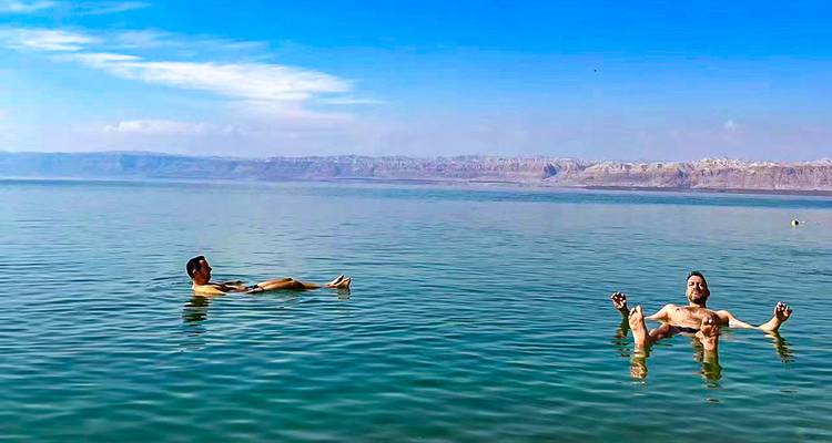 Two people floating in the Dead Sea with distant hills and a blue sky.