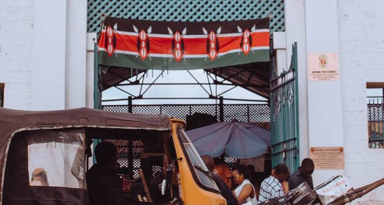 Entrée d'un marché avec un drapeau du Kenya au-dessus et un tuk-tuk garé à l'extérieur.