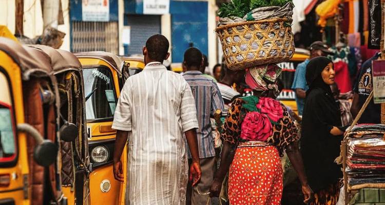 Rue animée avec des gens et des étals, femme portant un panier sur sa tête.