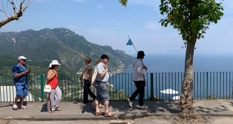 People walking by a railing overlooking a sea with mountains.