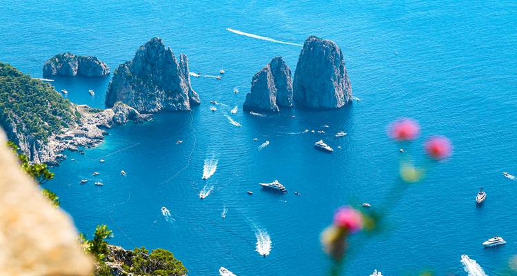 Aerial vista of Capri’s rugged coastline and Faraglioni stacks surrounded by yachts and speedboats on vivid blue water.