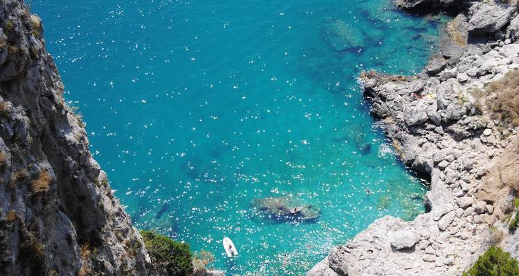 Sparkling turquoise Mediterranean water below steep rocky cliff with lone paddleboarder.