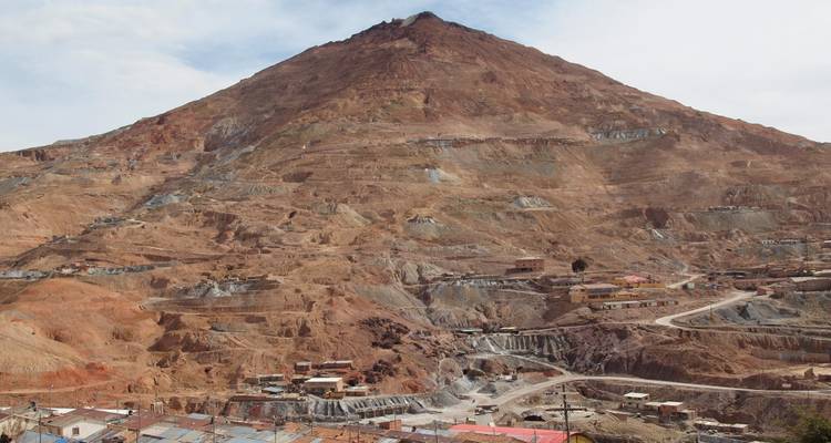 Ein massiver rotbraun gefärbter Berg, gezeichnet von Silberminen, ragt über eine raue Siedlung empor.