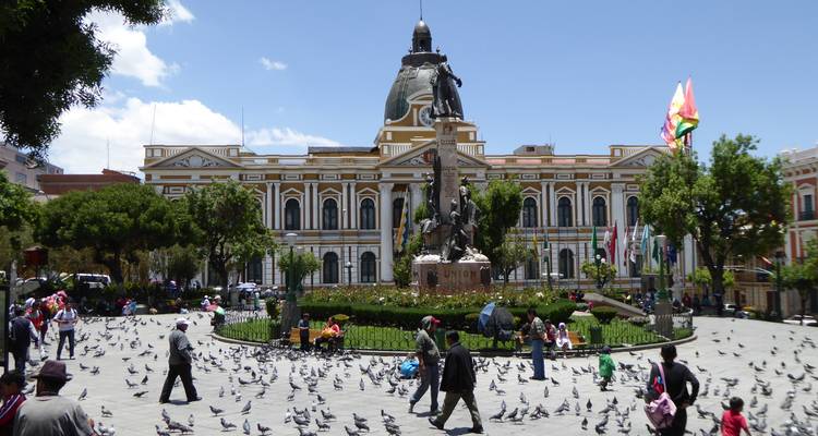 Belebter Platz mit Tauben, üppigen Gärten und Boliviens prunkvollem Regierungspalast im Hintergrund.