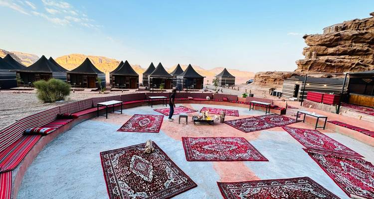 Kreisförmige Freiluft-Lounge mit gemusterten Teppichen in einem Wüstencamp, umrahmt von schwarzen Beduinenzelten und Sandsteinklippen