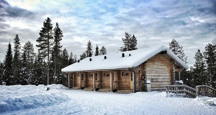 Gemütliche Holzhütte, bedeckt von tiefem Schnee, umgeben von hohen Kiefern unter einem dramatischen Winterhimmel.