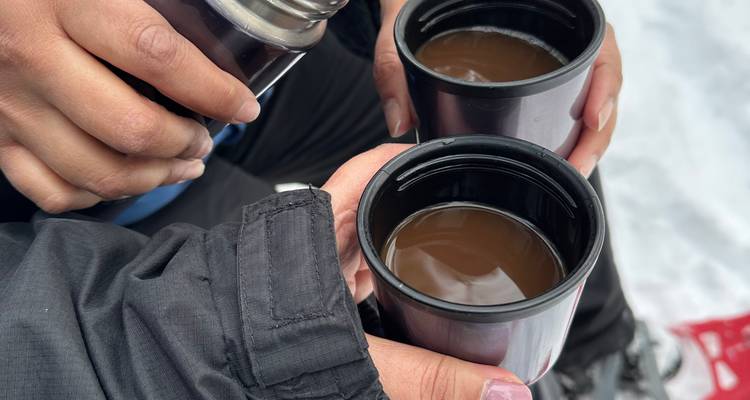 Zwei Personen wärmen ihre Hände an dampfenden Bechern mit heißem Getränk während einer verschneiten Outdoor-Pause.