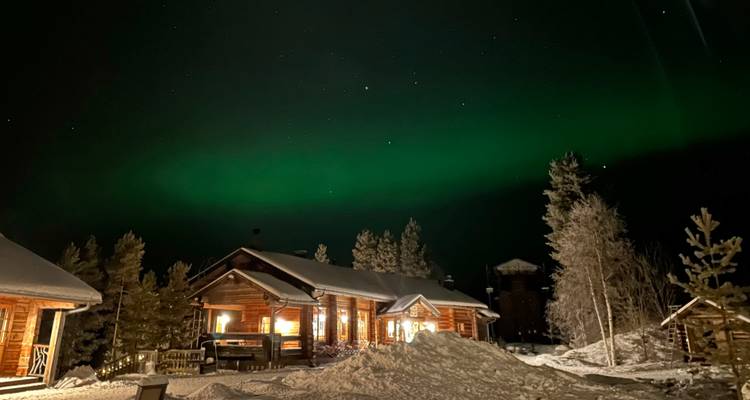 Das Nordlicht leuchtet grün über schneebedeckten Hütten in einer stillen Winternacht.