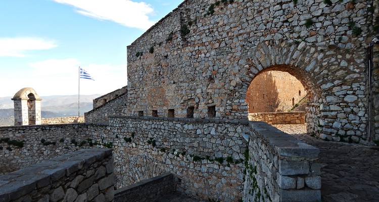 Des remparts de forteresse avec le drapeau grec surplombent des vallées lointaines et le littoral.