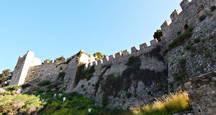 De hauts murs de château en pierre crénelés se dressent contre un ciel bleu dégagé.