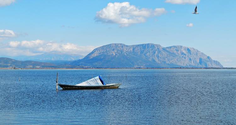 Un bateau en bois solitaire flotte sur les eaux tranquilles d'un lagon avec des montagnes et une mouette qui vole.