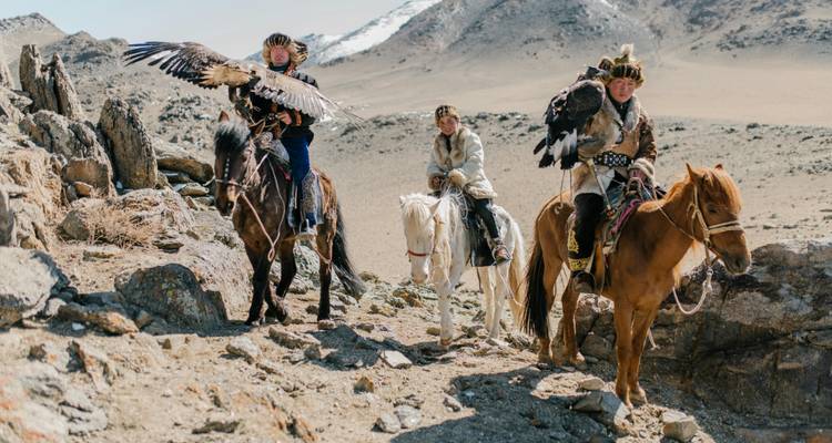 Tres personas a caballo sosteniendo águilas en un paisaje montañoso rocoso.