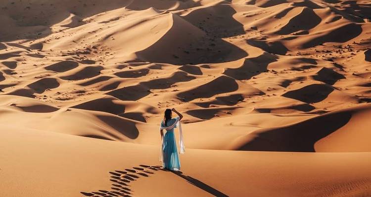 Personne en robe bleue marchant sur des dunes désertiques avec des empreintes de pas.