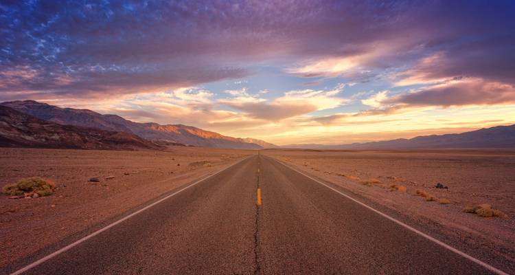 Route infinie qui disparaît dans un vaste paysage désertique sous des nuages de coucher de soleil dorés spectaculaires.