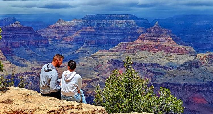 Couple assis sur le bord d'une falaise profitant de vues panoramiques et étagées du Grand Canyon sous un ciel chargé.