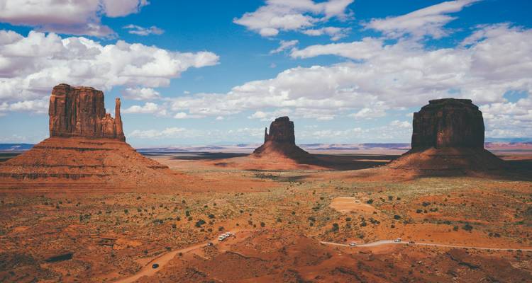 Trois imposants pitons de grès se dressent isolés dans le vaste désert rouge de Monument Valley sous un ciel bleu.