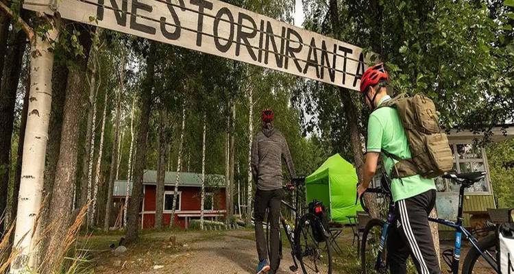 Des cyclistes entrent dans une zone avec un panneau en bois et des cabanes.