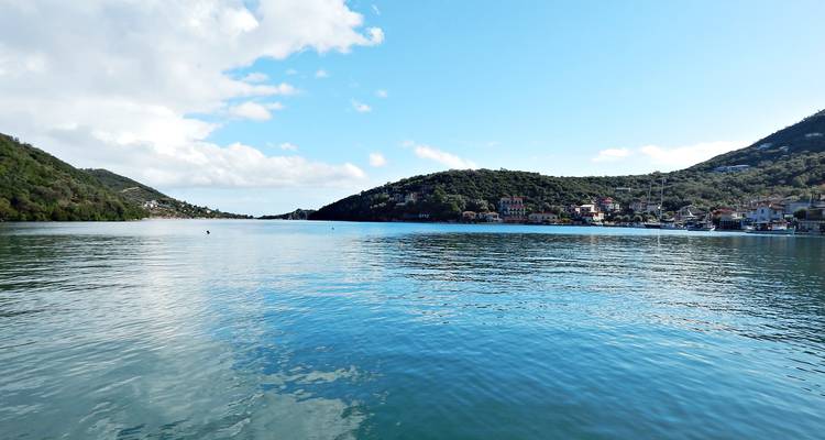 Baie calme et abritée avec petit village et collines vertes reflétées sur l'eau bleue immobile sous la lumière vive du jour.