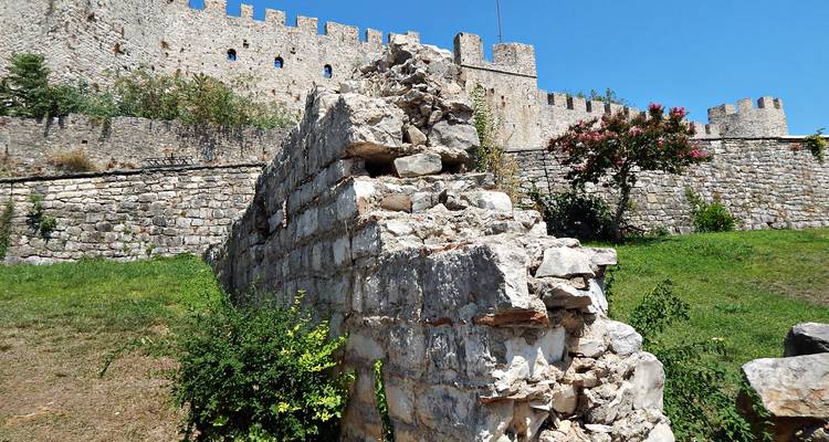 Mur de pierre en ruine menant à un château médiéval fortifié avec des tours crénelées sous un ciel bleu dégagé.