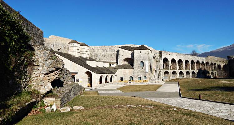 Complexe de forteresse en pierre avec des arches et une petite église à dôme contre un ciel bleu dégagé.