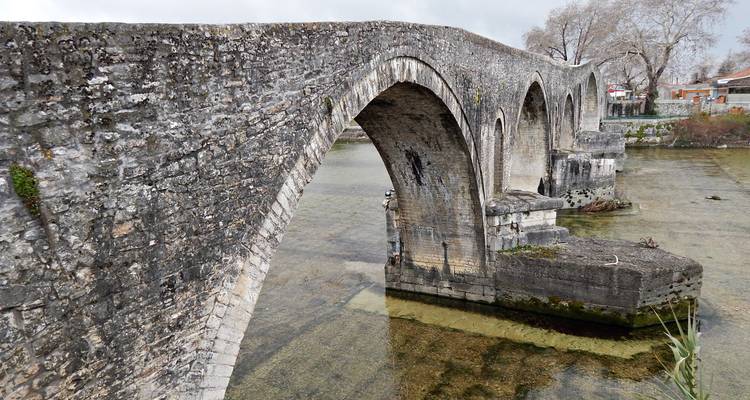 Puente de arco de piedra atraviesa un río poco profundo con agua transparente verdosa.