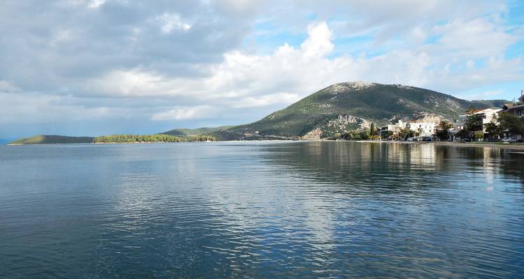 Kalme kustwateren die een gedeeltelijk bewolkte lucht weerkaatsen met groene heuvels en een kustdorp in de verte.