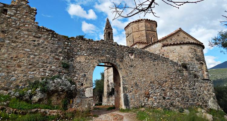 Stenen booggang die leidt naar een klein middeleeuws kerkcomplex met blauwe lucht en wolken.