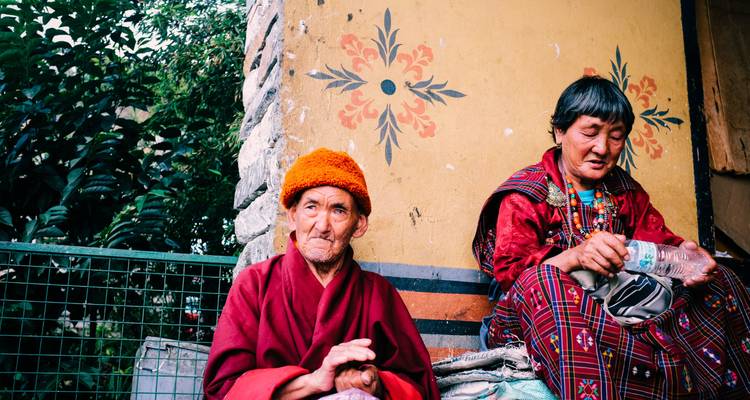 Dos personas mayores con vestimenta tradicional butanesa sentadas junto a una pared.