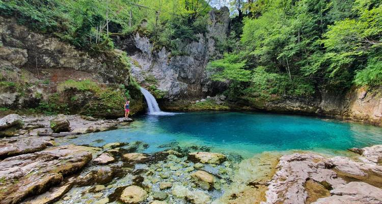 Persona de pie cerca de una pequeña cascada y agua azul en una zona rocosa.
