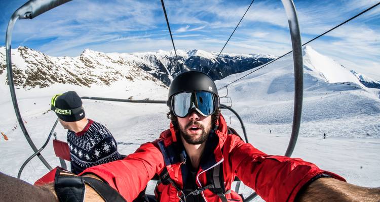 Selfie of two people on a ski lift with snowy peaks.
