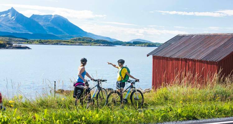 Deux cyclistes s'arrêtent près d'un cabanon rouge dominant un fjord pittoresque avec des montagnes en arrière-plan.