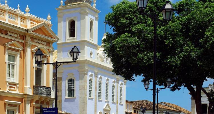 Fachada colorida de iglesia de la época colonial con campanario ornamentado enmarcado por farolas vintage y árboles frondosos contra un cielo brillante.