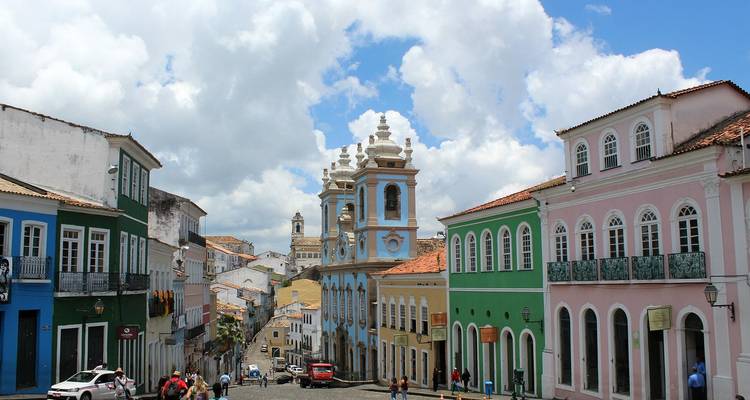 Calle empedrada bordeada de casas coloniales pintadas en colores pastel bullendo de peatones bajo un cielo de nubes ondulantes.