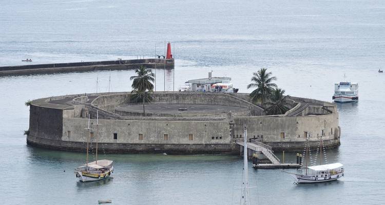 Una fortaleza marina circular única se alza desde las tranquilas aguas del puerto, rodeada de pequeñas embarcaciones y palmeras.