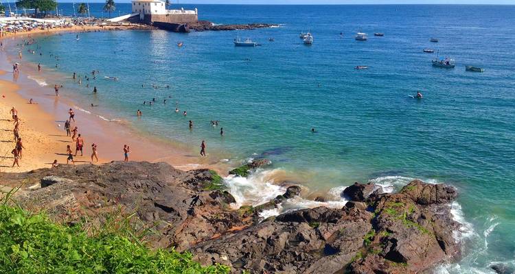 Cala de playa concurrida con nadadores en agua turquesa, primer plano rocoso y un pequeño fuerte visible en la distancia.