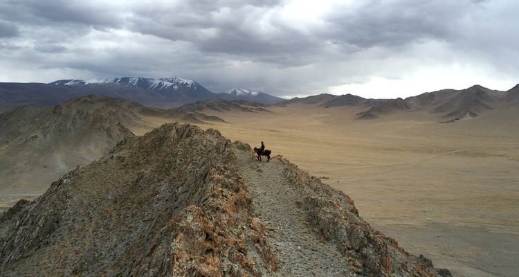 Jinete solitario y caballo en una cresta rocosa de montaña contra un cielo nublado.