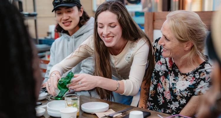 Viajeros compartiendo bebidas y platillos coreanos en una mesa al aire libre, riendo juntos