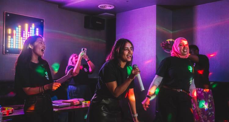 Los viajeros cantan con entusiasmo en una sala de karaoke iluminada con neón, sosteniendo micrófonos y teléfonos