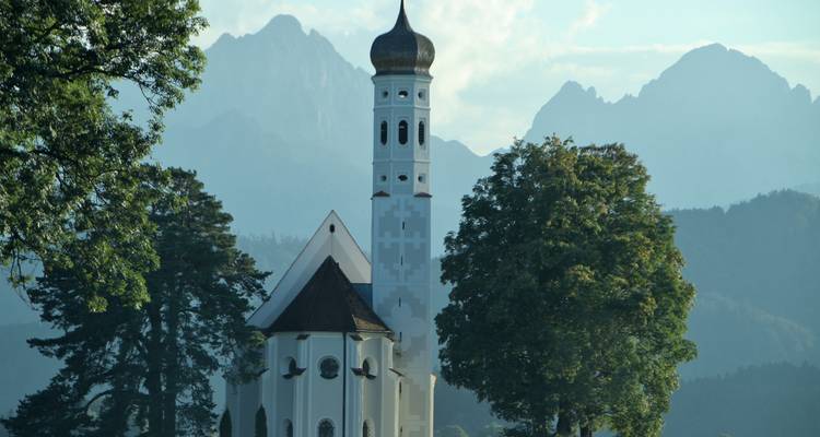 Eine Kirche eingebettet zwischen Bäumen mit Bergen im Hintergrund.