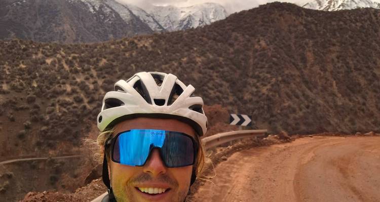 Radfahrer, der ein Selfie auf einer unbefestigten Bergstraße mit schneebedeckten Gipfeln und Wüstenhügeln im Hintergrund macht