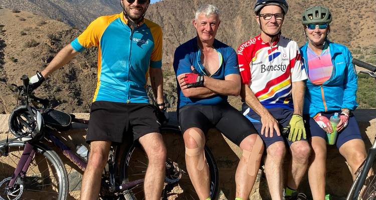 Vier Radfahrer ruhen sich auf einer Steinmauer aus, die über die raue Berglandschaft Marokkos blickt