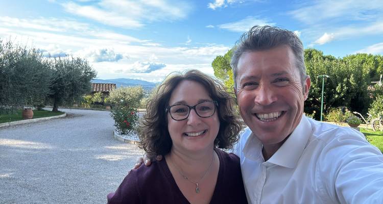 Dos personas sonrientes posando al aire libre contra un fondo escénico con árboles y un sendero de grava.