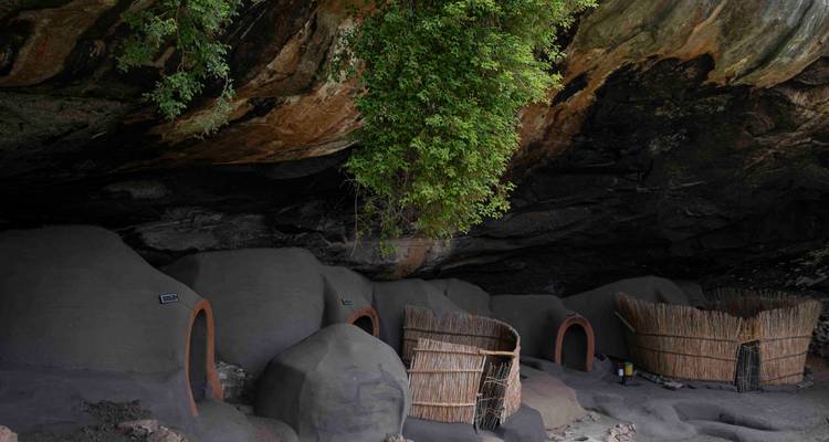 Traditionele modderhutten onder een rotsrichel omgeven door natuurlijke begroeiing.